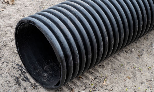 A large black corrugated pipe lies horizontally on sandy ground, with textured ridges visible. The setting appears outdoors, conveying an industrial tone.